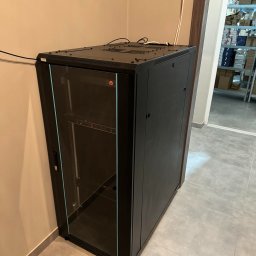 INVICTEL PIKUŁA SPÓŁKA JAWNA - Czarna szafa rack z przeszklonymi drzwiami, widoczny wyświetlacz temperatury, kable na górze, magazyn w tle. Szafa stoi w rogu pomieszczenia na szarych płytkach.