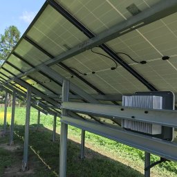 INVICTEL PIKUŁA SPÓŁKA JAWNA - Instalacja paneli słonecznych od spodu, widoczne mocowania i inwerter w Kamyku. Metalowa konstrukcja na tle zieleni i błękitnego nieba.