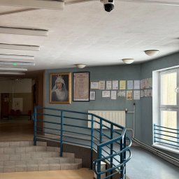 INVICTEL PIKUŁA SPÓŁKA JAWNA - Kamera monitoringu na suficie w budynku z obrazem i dyplomami na ścianie. Widok na schody i korytarz.