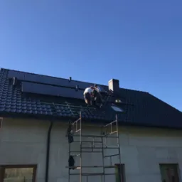 Montaż paneli słonecznych na dachu domu z czarną dachówką. Dwóch monterów pracuje na wysokości, widoczne rusztowanie i błękitne niebo.