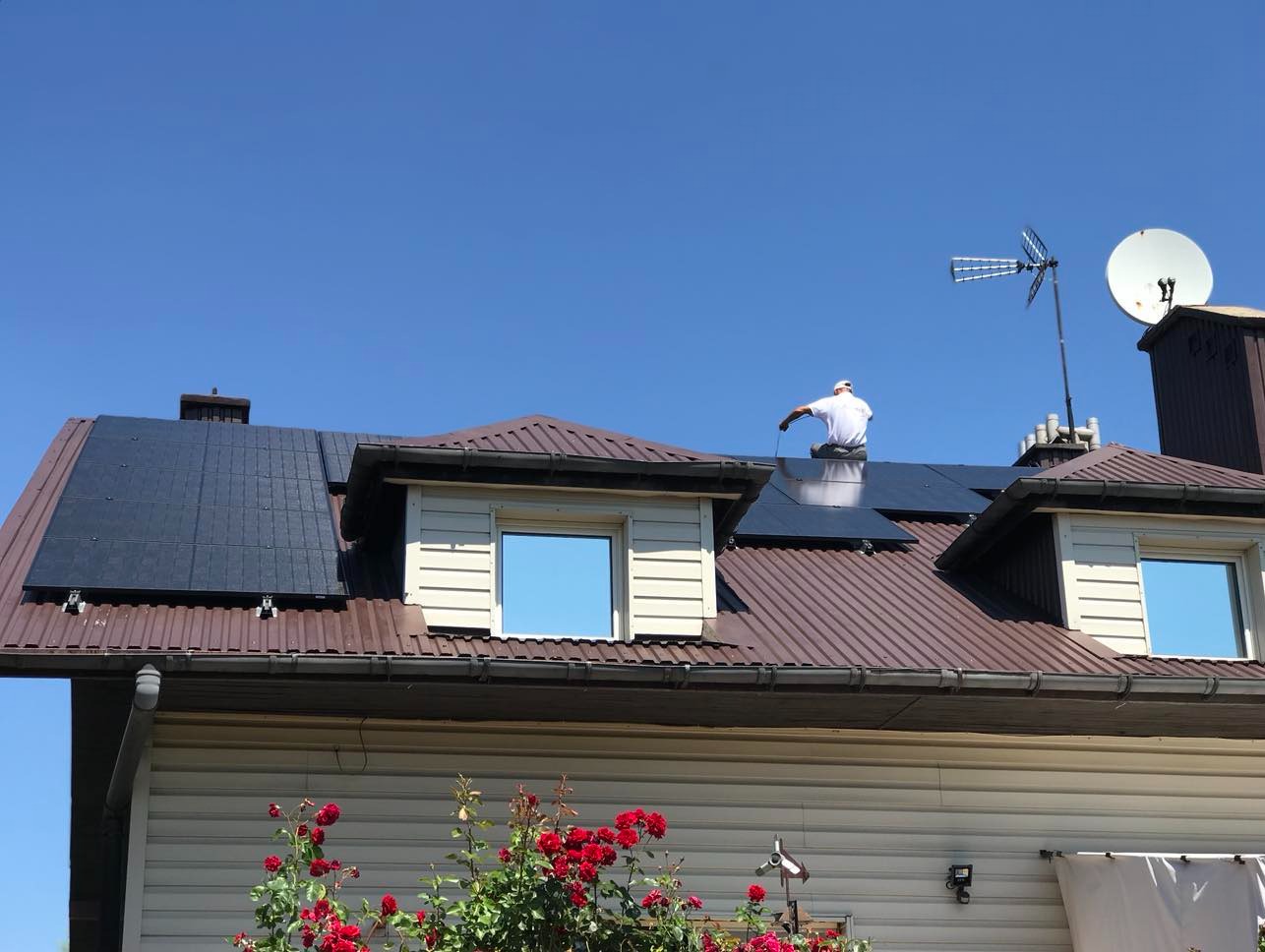 Instalacja paneli słonecznych na brązowym dachu domu z anteną satelitarną i telewizyjną. Pracownik w białej koszulce łączy przewody na panelach.
