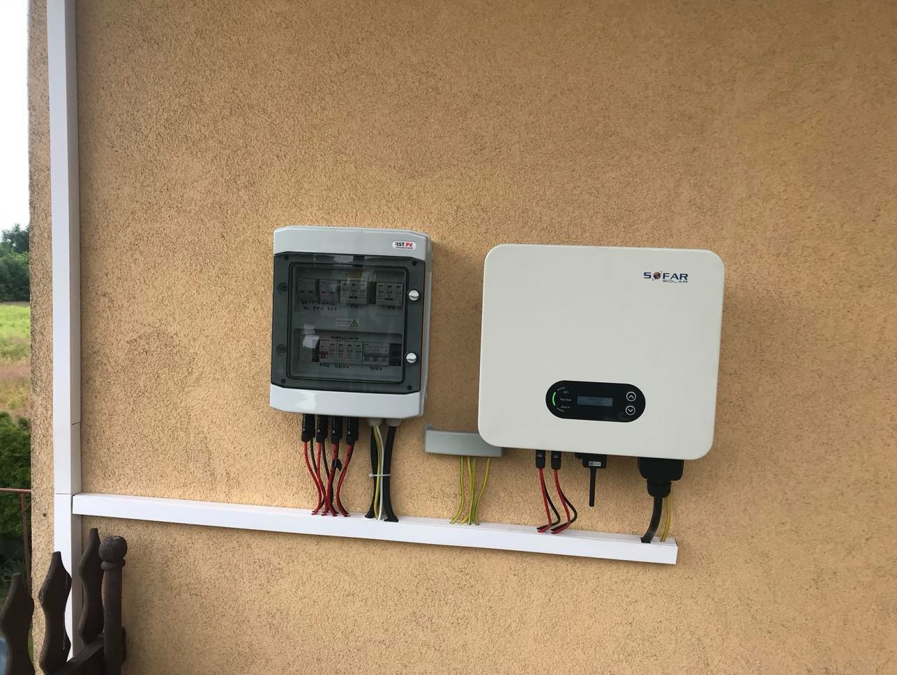 Biały inwerter SOFAR i skrzynka elektryczna z przewodami na beżowej ścianie budynku, instalacja fotowoltaiczna na zewnątrz, widok z dołu.