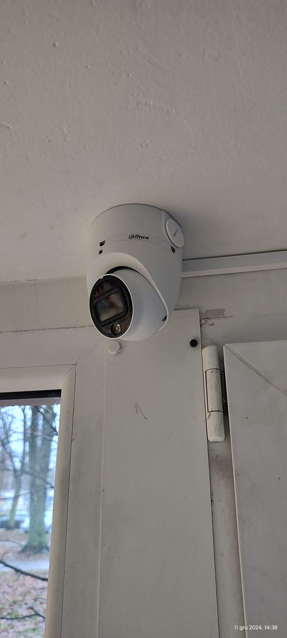 Kopułkowa kamera Dahua zamontowana przy suficie, widok z dołu. Obok fragment okna z widokiem na drzewa i drzwi. Białe ściany z widocznymi śladami użytkowania.