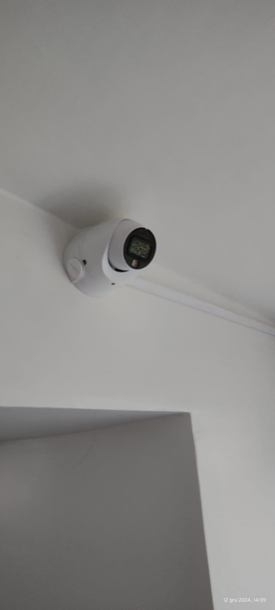 Kamera monitoringu w kształcie kuli, zamontowana na białym suficie w rogu pomieszczenia. Widoczny przewód. Ujęcie z dołu pod kątem.