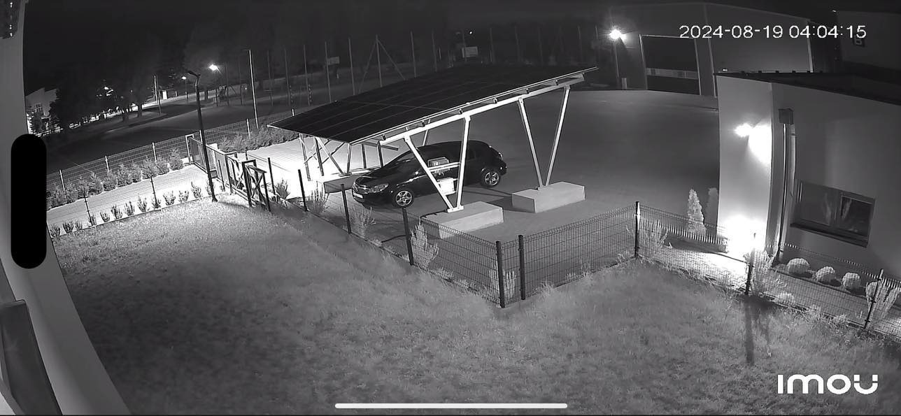Nocny widok z kamery monitoringu na samochód pod wiatą solarną. Widoczny budynek, ogrodzenie i trawnik. Data i godzina w prawym górnym rogu.