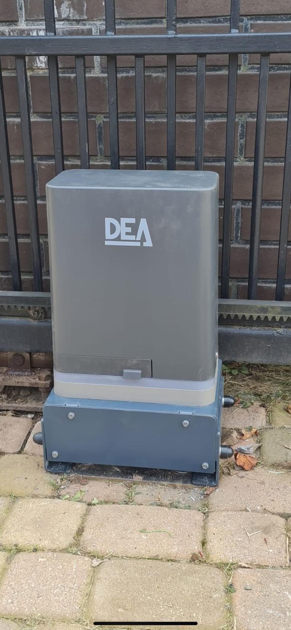 Szary automat do bram DEA zamontowany na kostce brukowej przy metalowym ogrodzeniu z ceglanym murem w tle. Widoczne śruby mocujące i przewody.