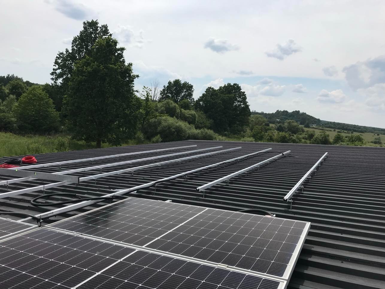 Instalacja paneli słonecznych na dachu z blachy trapezowej. Widoczne szyny montażowe i częściowo ułożone panele. Tło: zielone drzewa i błękitne niebo z chmurami.