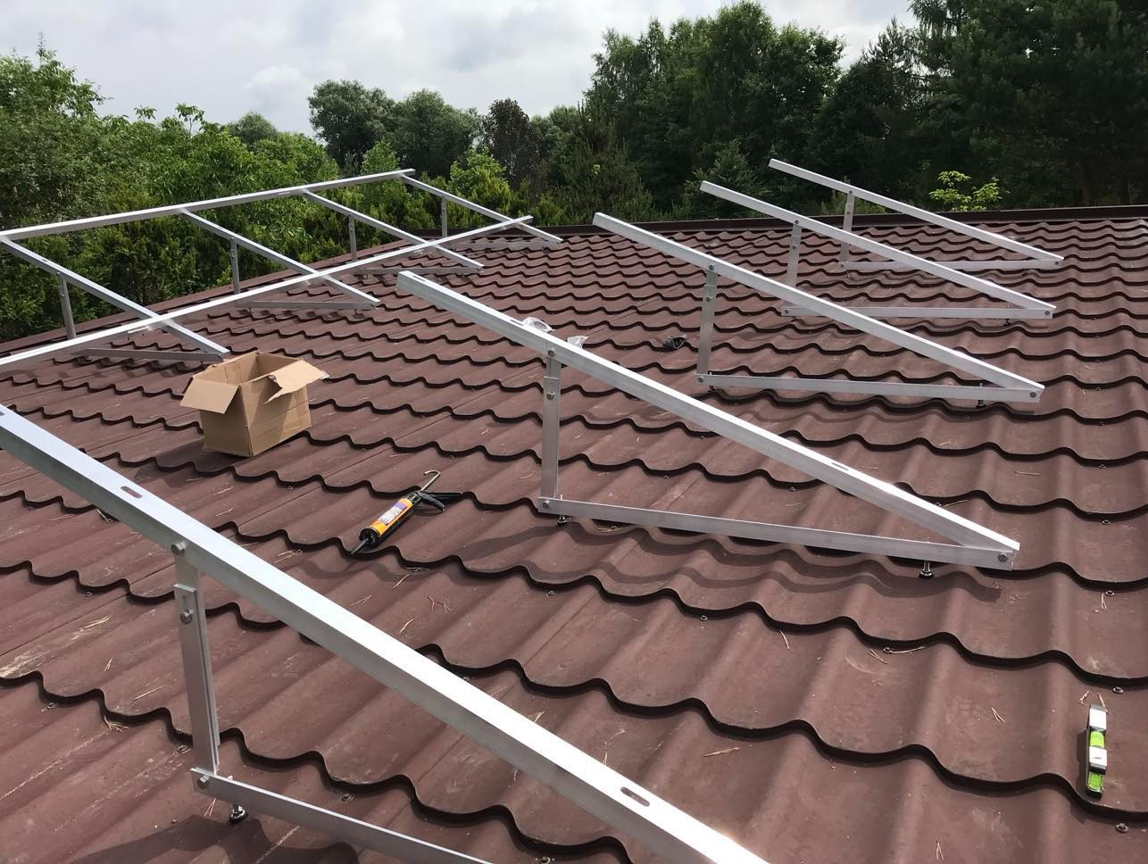 Montaż konstrukcji wsporczej pod panele fotowoltaiczne na brązowym dachu z blachodachówki. Widoczny karton, tuba silikonu i poziomica. Tło: zielone drzewa.