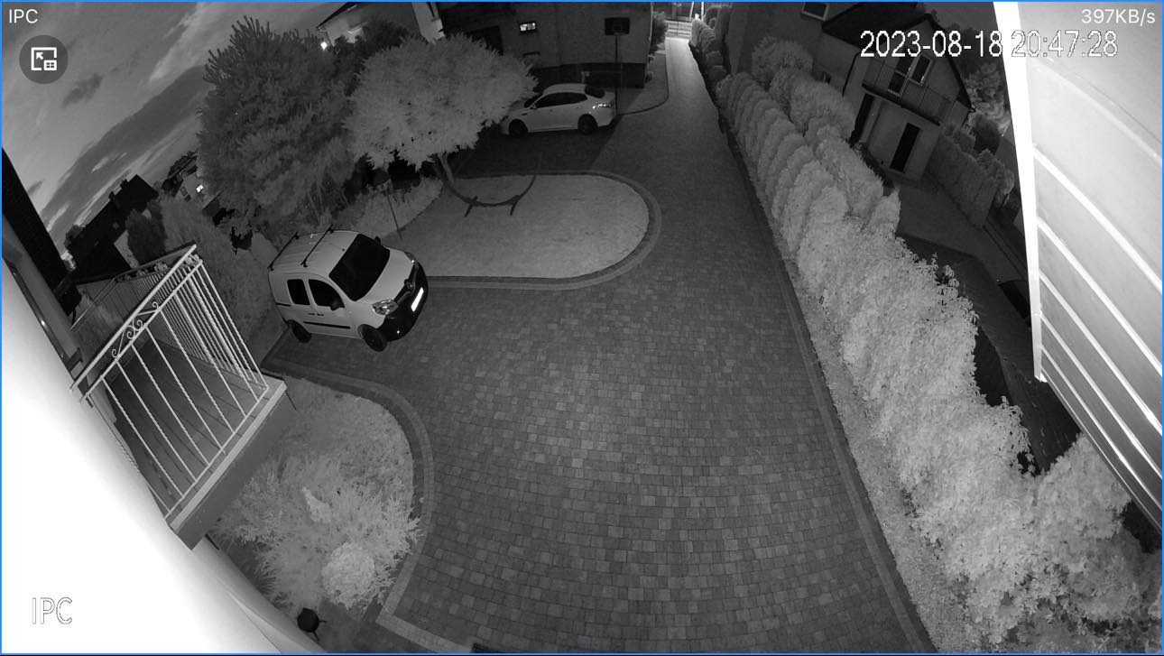 Nocny widok z kamery monitoringu: biały samochód dostawczy na brukowanym podjeździe, drugi samochód w tle, drzewa i krzewy, budynek z balkonem.