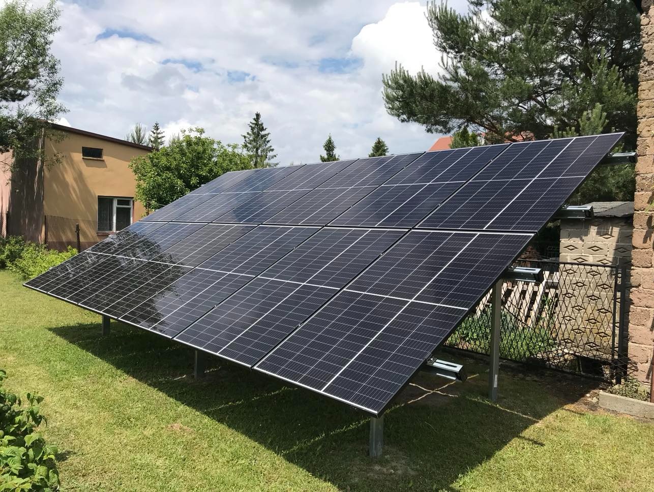 Panele fotowoltaiczne zamontowane na metalowej konstrukcji na trawniku, z budynkiem i drzewami w tle, w słoneczny dzień. Widok z bliska na gotową instalację.