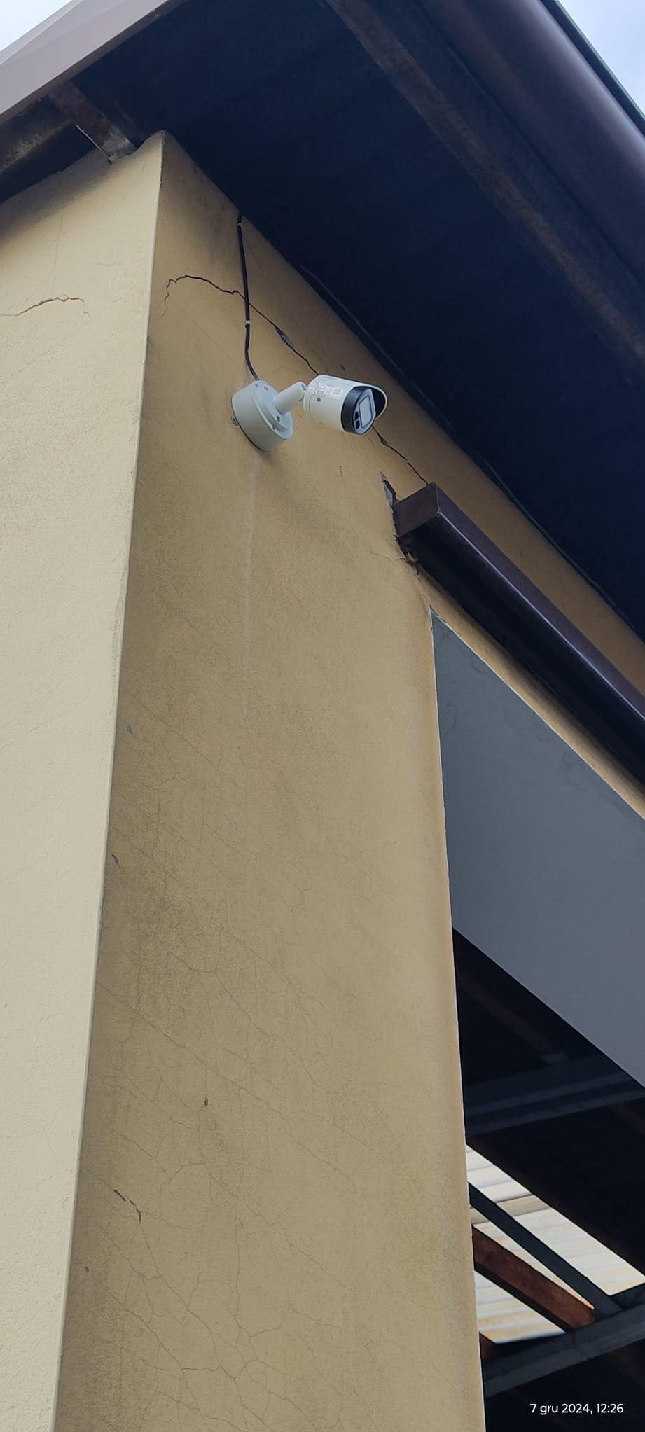 Zamontowana kamera monitoringu na rogu budynku z beżową elewacją, widoczne pęknięcia na ścianie. Kabel kamery prowadzony wzdłuż ściany pod okapem dachu.
