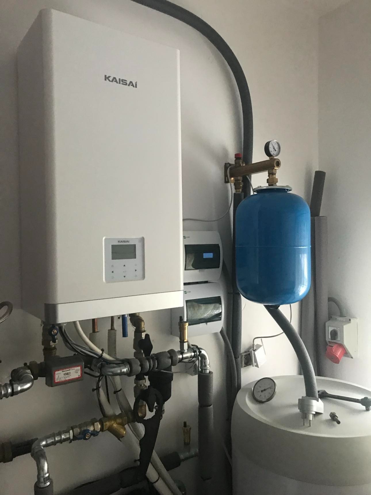 Biały piec gazowy marki Kaisai z podłączeniami hydraulicznymi, zbiornikiem wyrównawczym i elementami instalacji w pomieszczeniu technicznym. Widoczne przewody i zawory.