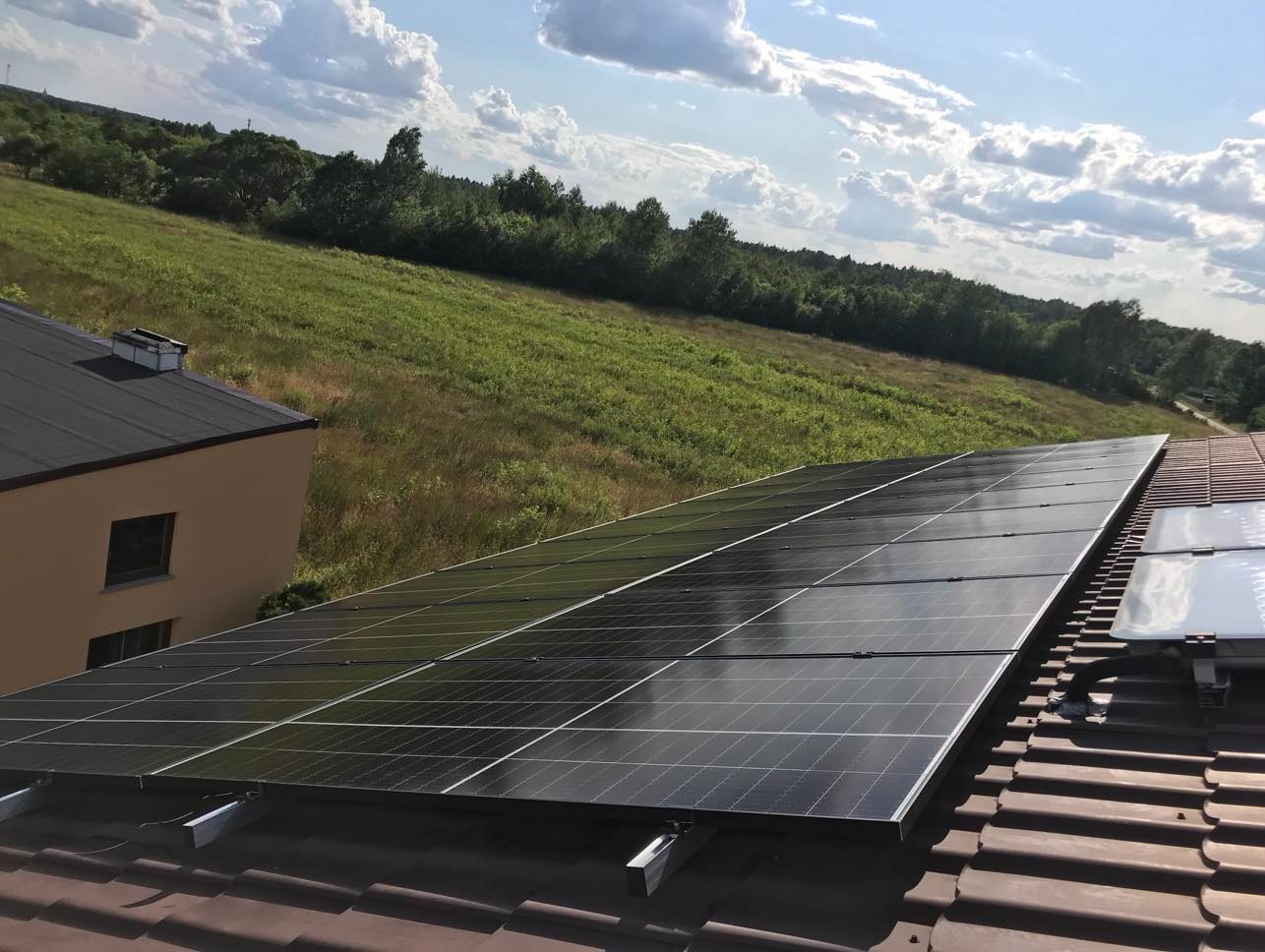 Panele słoneczne na dachu domu z brązową dachówką, w tle zielone wzgórze i błękitne niebo z chmurami. Widok z góry, jasne oświetlenie.