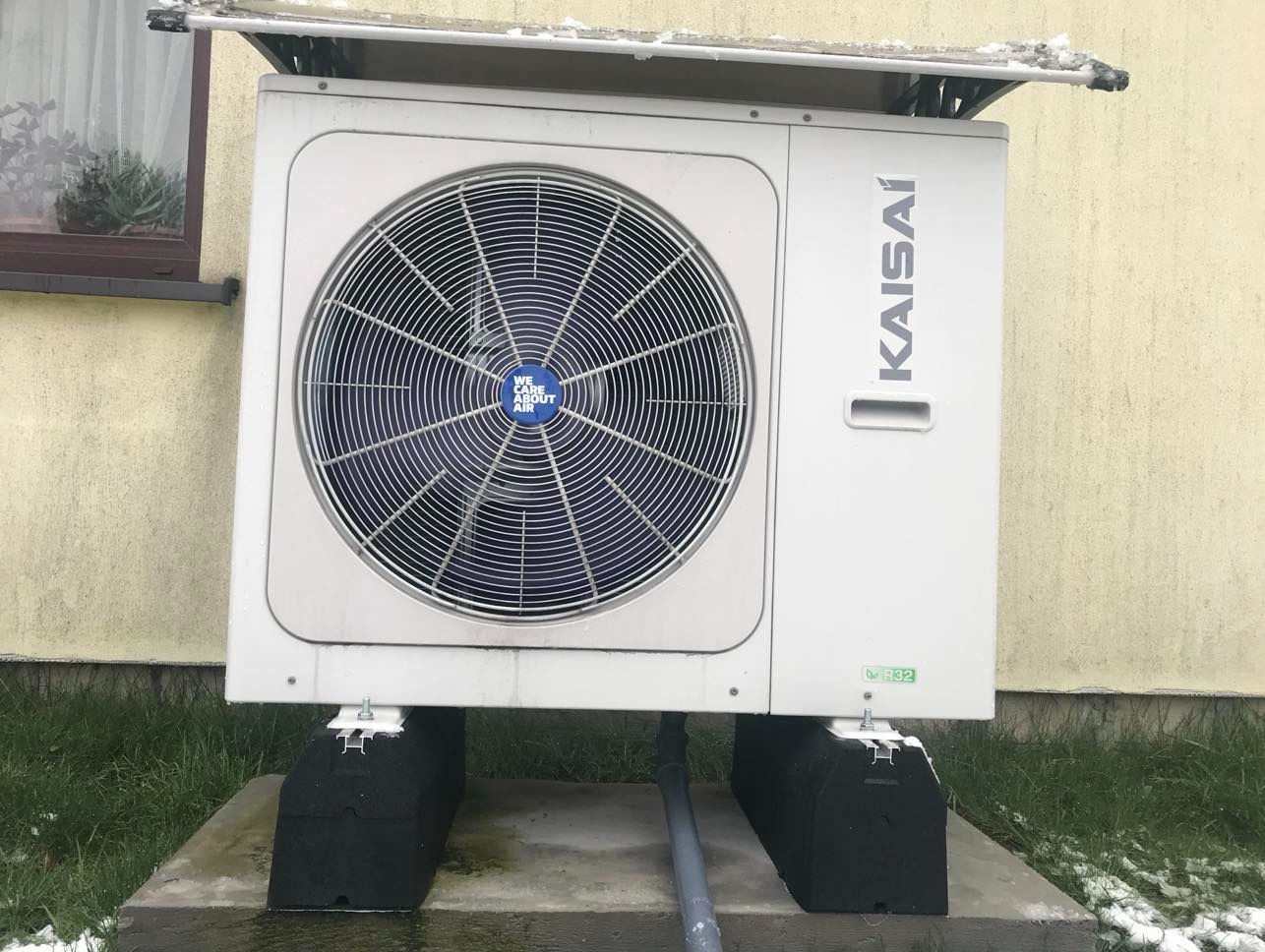 Klimatyzator Kaisai zamontowany na zewnątrz budynku, na betonowej podstawie. Widoczne elementy instalacji i fragment trawnika. Ściana budynku w tle.