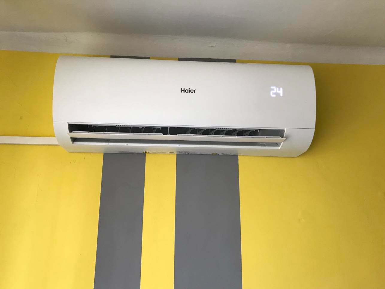 Biały klimatyzator Haier z wyświetlaczem 24°C zamontowany na żółto-szarej ścianie z pionowymi pasami. Widok z przodu, oświetlenie dzienne.