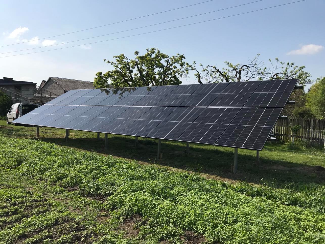 Instalacja paneli słonecznych na trawiastym terenie, widok z boku, z domem i samochodem w tle. Panele zamontowane na metalowych wspornikach.