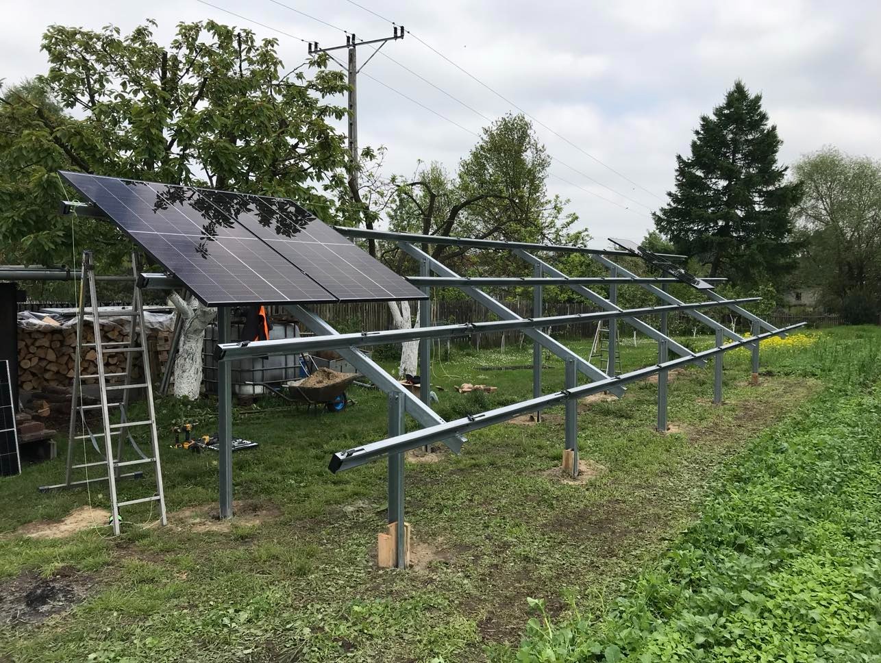 Montaż paneli słonecznych na metalowej konstrukcji w ogrodzie, widoczne narzędzia i drabina. Częściowo zainstalowane panele, w tle drzewa i słupy energetyczne.