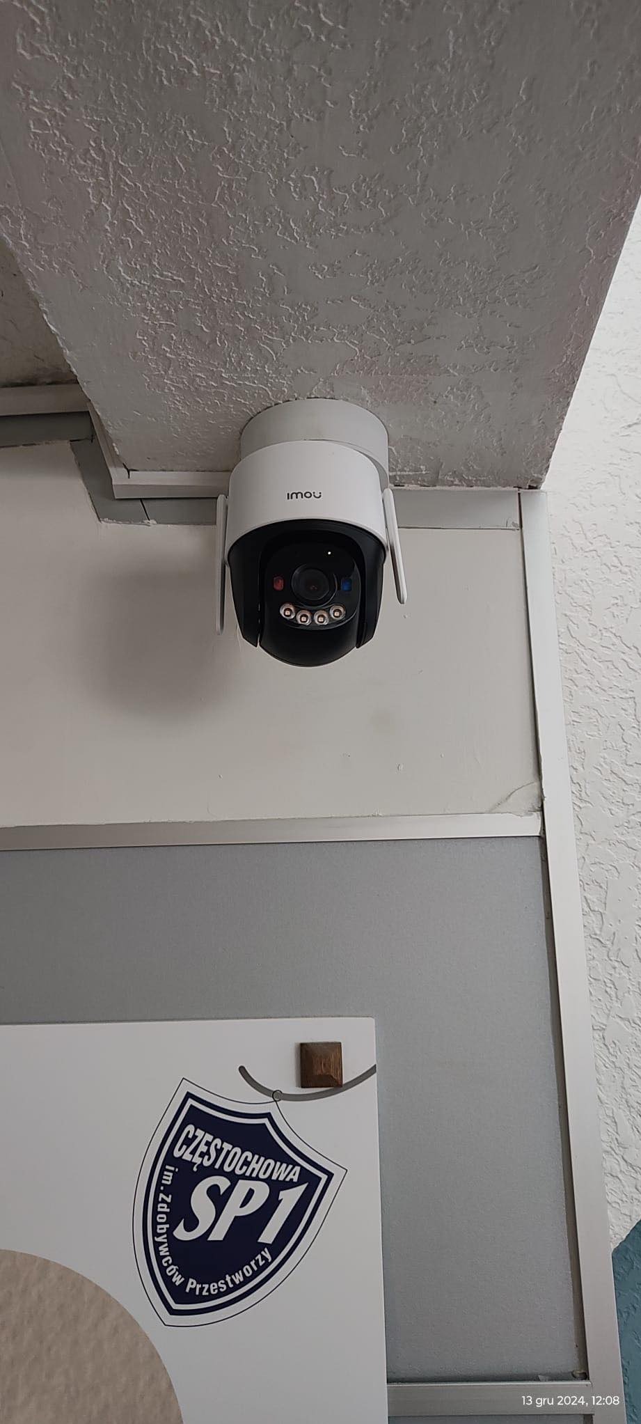 Kamera monitoringu Imou, zamontowana pod sufitem, w rogu pomieszczenia. Widoczny fragment ściany z tablicą informacyjną. Biała obudowa kamery kontrastuje z fakturą sufitu.