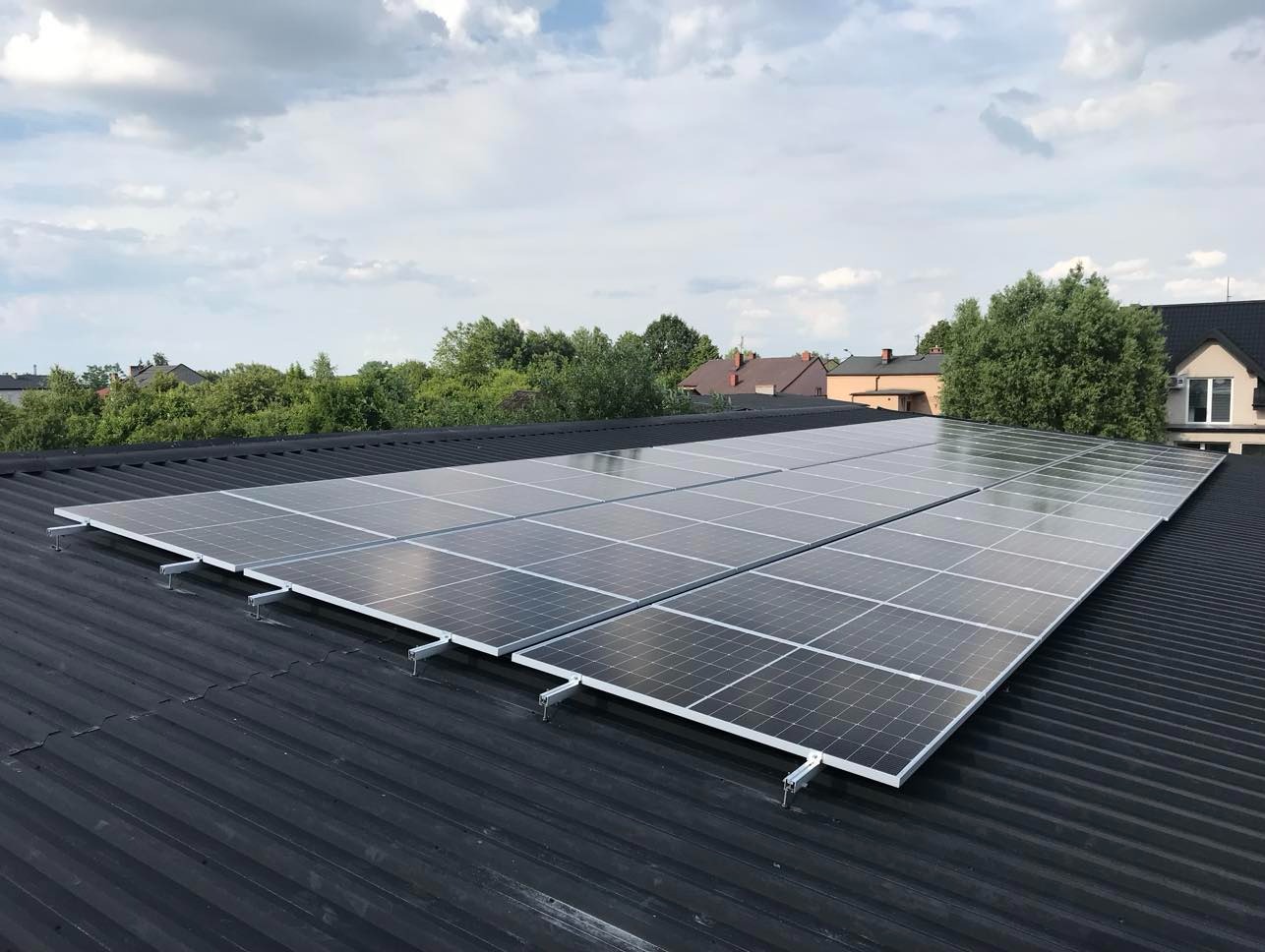 Panele słoneczne zamontowane na czarnym, blaszanym dachu domu jednorodzinnego. Widok z góry, w tle zieleń drzew i błękitne niebo z chmurami.