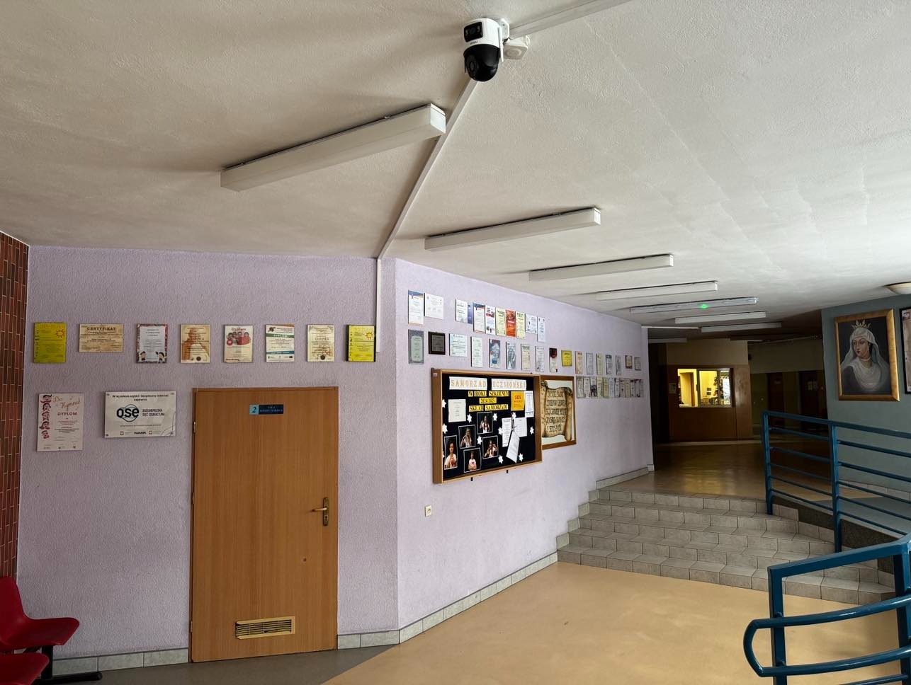 Kamera monitoringu kopułkowa zamontowana na suficie w korytarzu budynku. Na ścianach wiszą certyfikaty i tablice informacyjne, oświetlenie jarzeniowe.