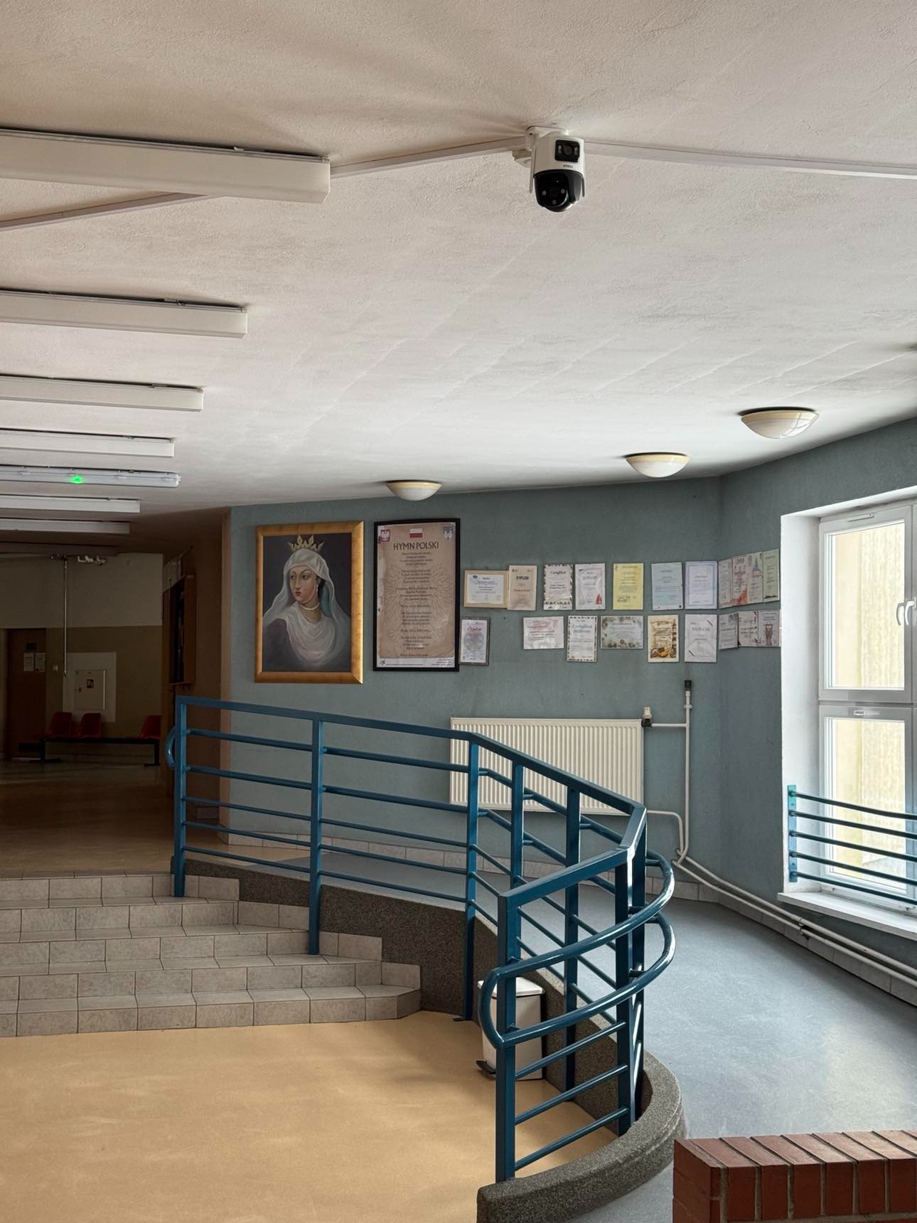 Kamera monitoringu na suficie w budynku z obrazem i dyplomami na ścianie. Widok na schody i korytarz.