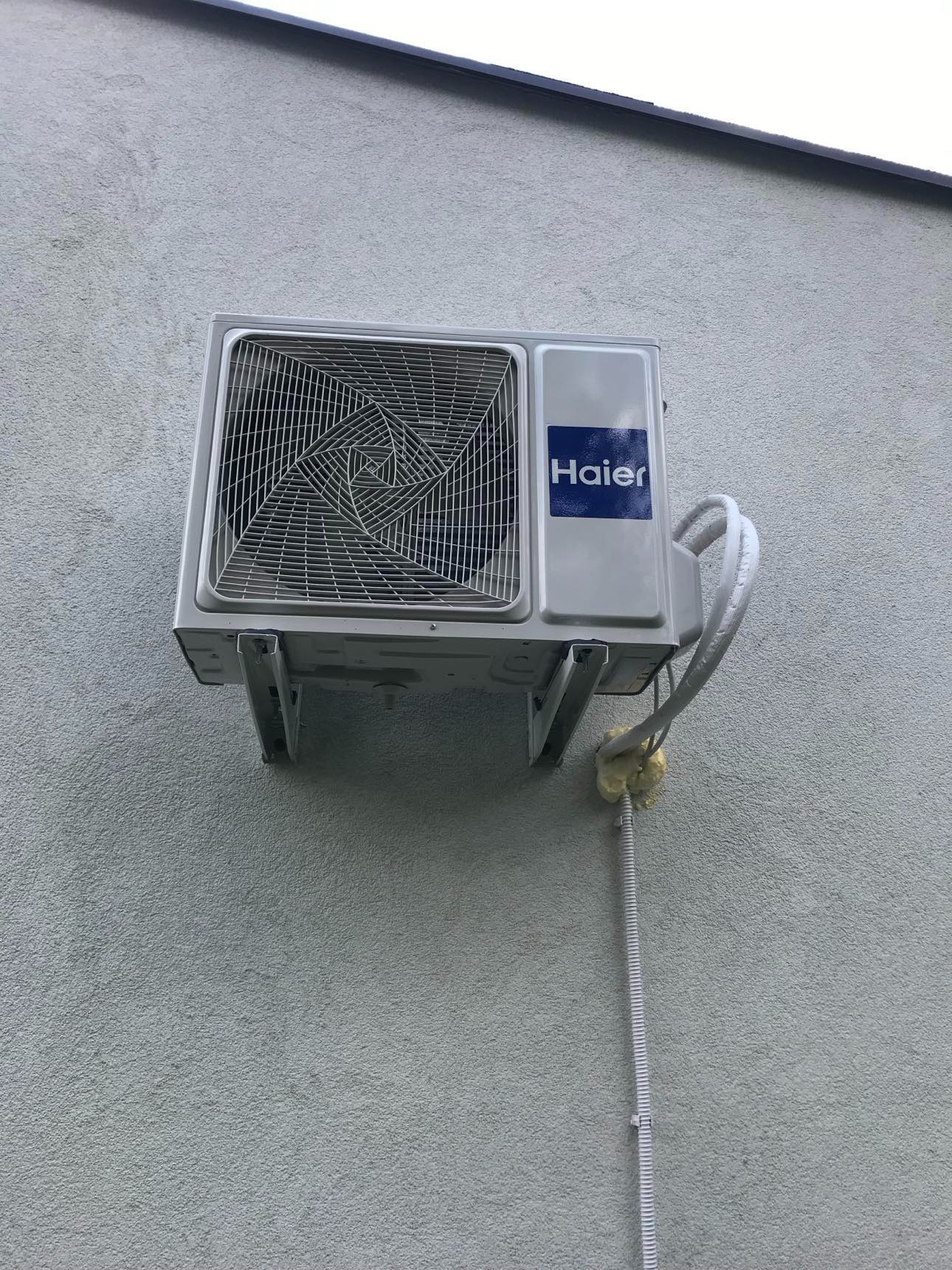 Zamontowana na szarej ścianie zewnętrzna jednostka klimatyzacji Haier z widocznymi przewodami i pianką montażową. Ujęcie z dołu, lekko z lewej strony.