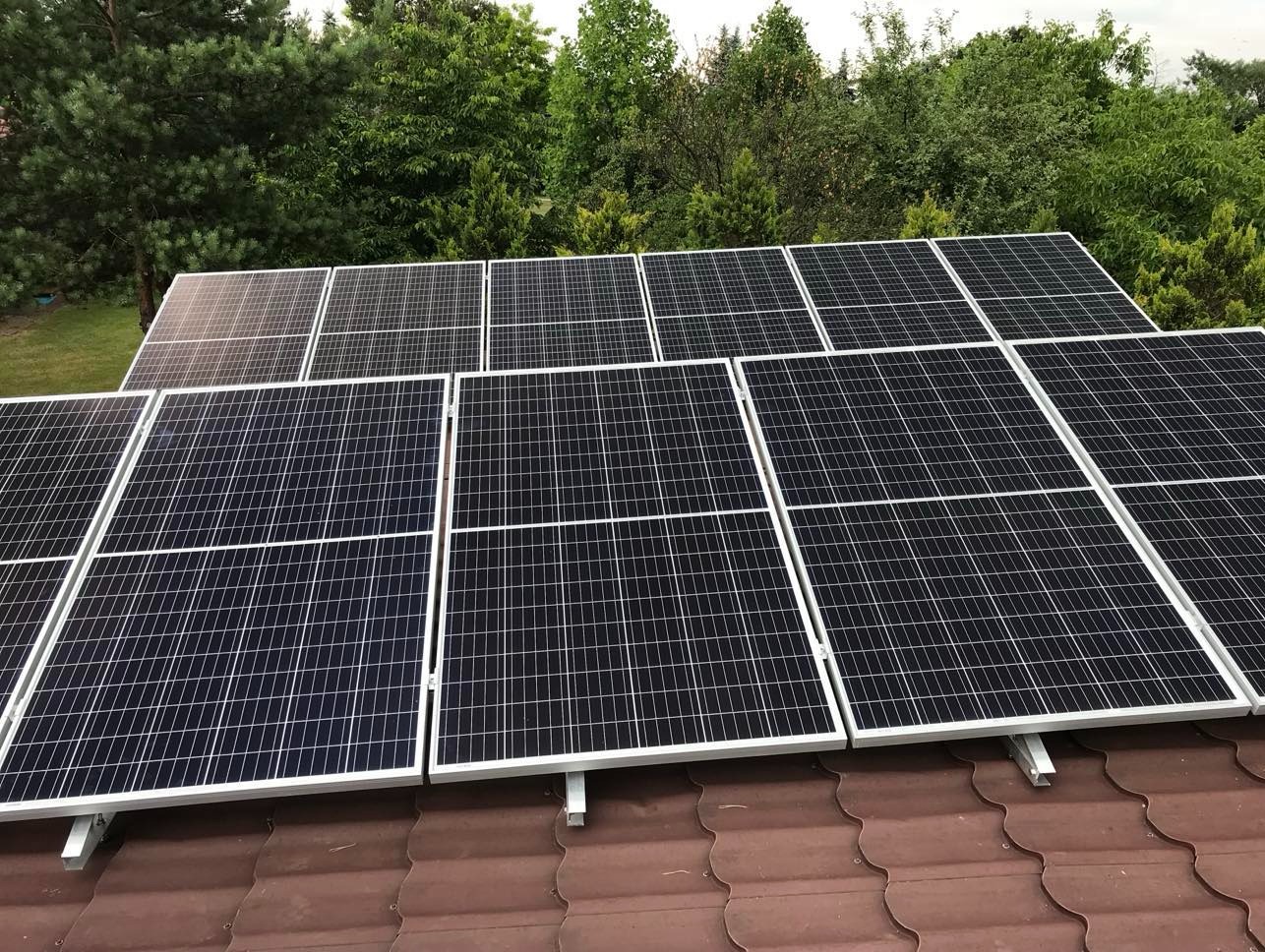 Panele słoneczne na brązowym dachu z dachówki, otoczone zielenią drzew. Widok z góry ukazuje symetryczny układ paneli i ich srebrne ramy.