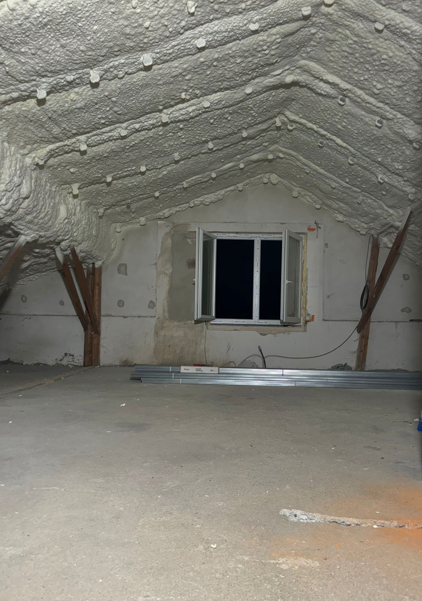 Poddasze po ociepleniu pianką PUR z widocznym oknem i elementami konstrukcyjnymi. Biała, nierówna struktura pianki pokrywa całą powierzchnię dachu.