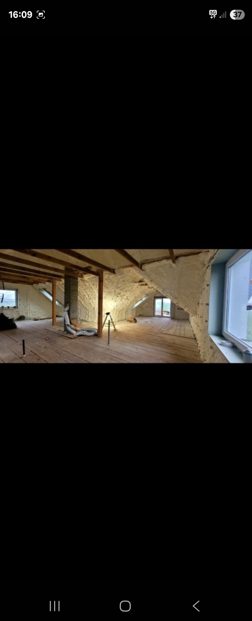 Poddasze z drewnianą podłogą po aplikacji izolacji piankowej na ścianach i suficie, widoczne okno i elementy konstrukcyjne.