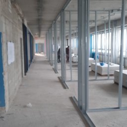 Medbud - Montaż metalowej konstrukcji ścian działowych w długim korytarzu. Widoczne profile, płyty karton-gips i pracownicy. Surowy stan wykończenia.