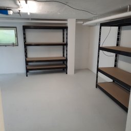 Medbud - Białe, świeżo pomalowane pomieszczenie z metalowymi regałami z brązowymi półkami. Widoczne rury pod sufitem i okno. Minimalizm i czystość.