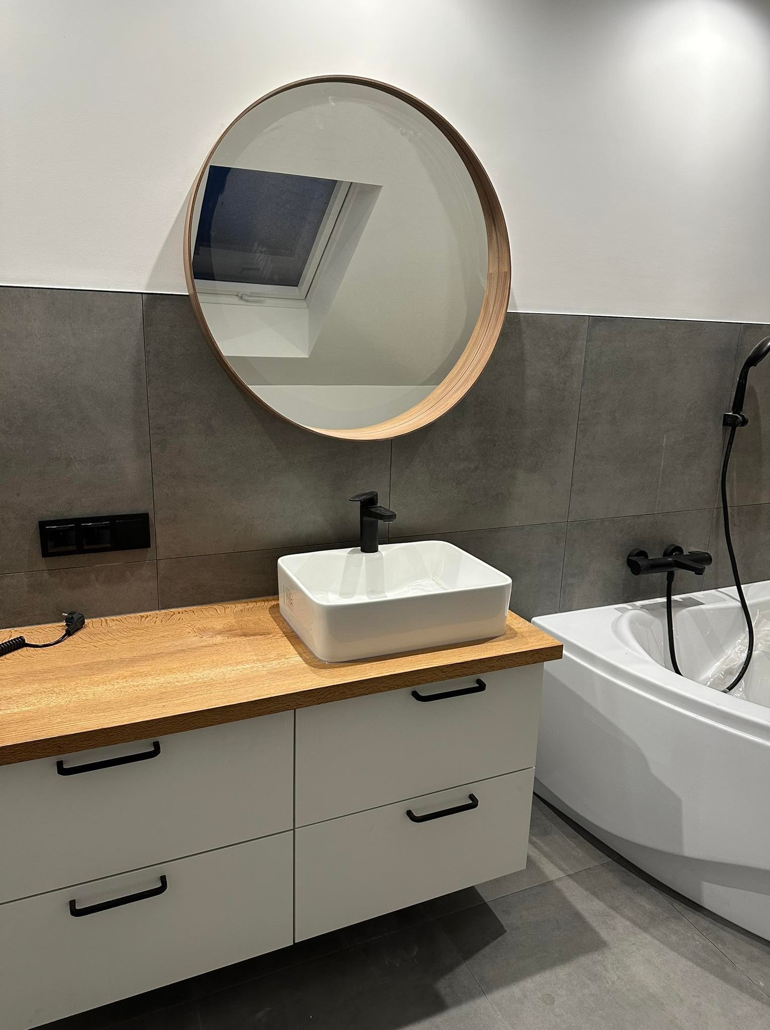 Nowoczesna łazienka z drewnianym blatem, białą szafką i umywalką nablatową. Okrągłe lustro z drewnianą ramą odbija okno dachowe.