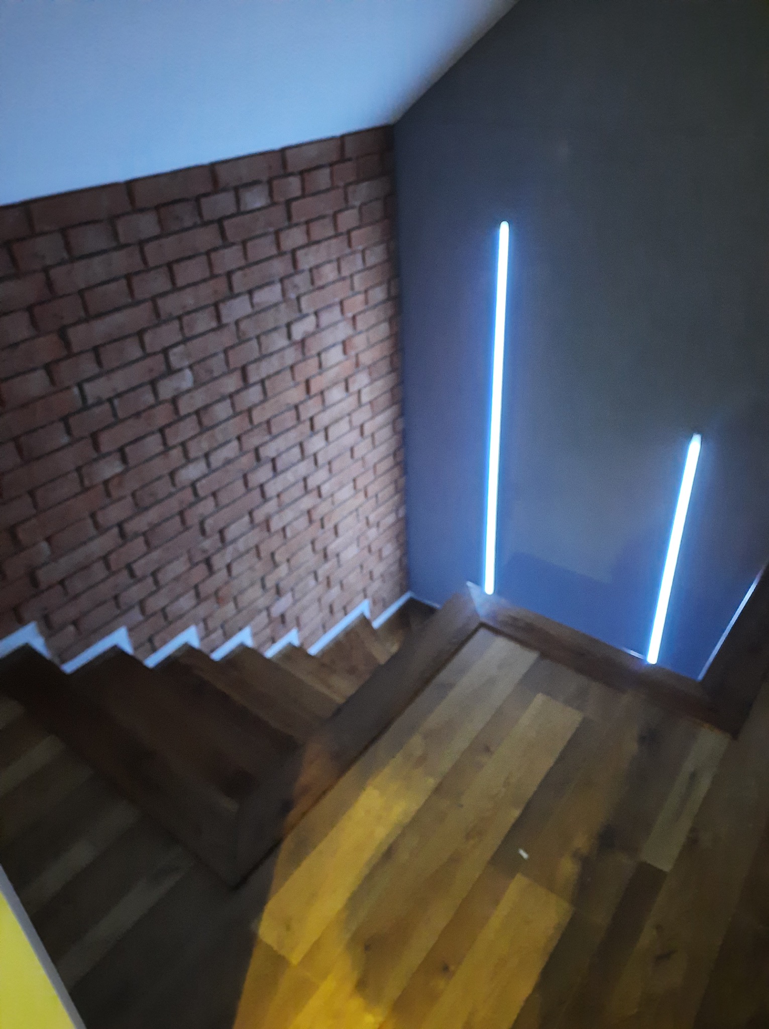 Schody z drewnianymi stopniami, ceglana ściana i pionowe, niebieskie oświetlenie LED. Nowoczesne wykończenie wnętrza, detale architektoniczne.