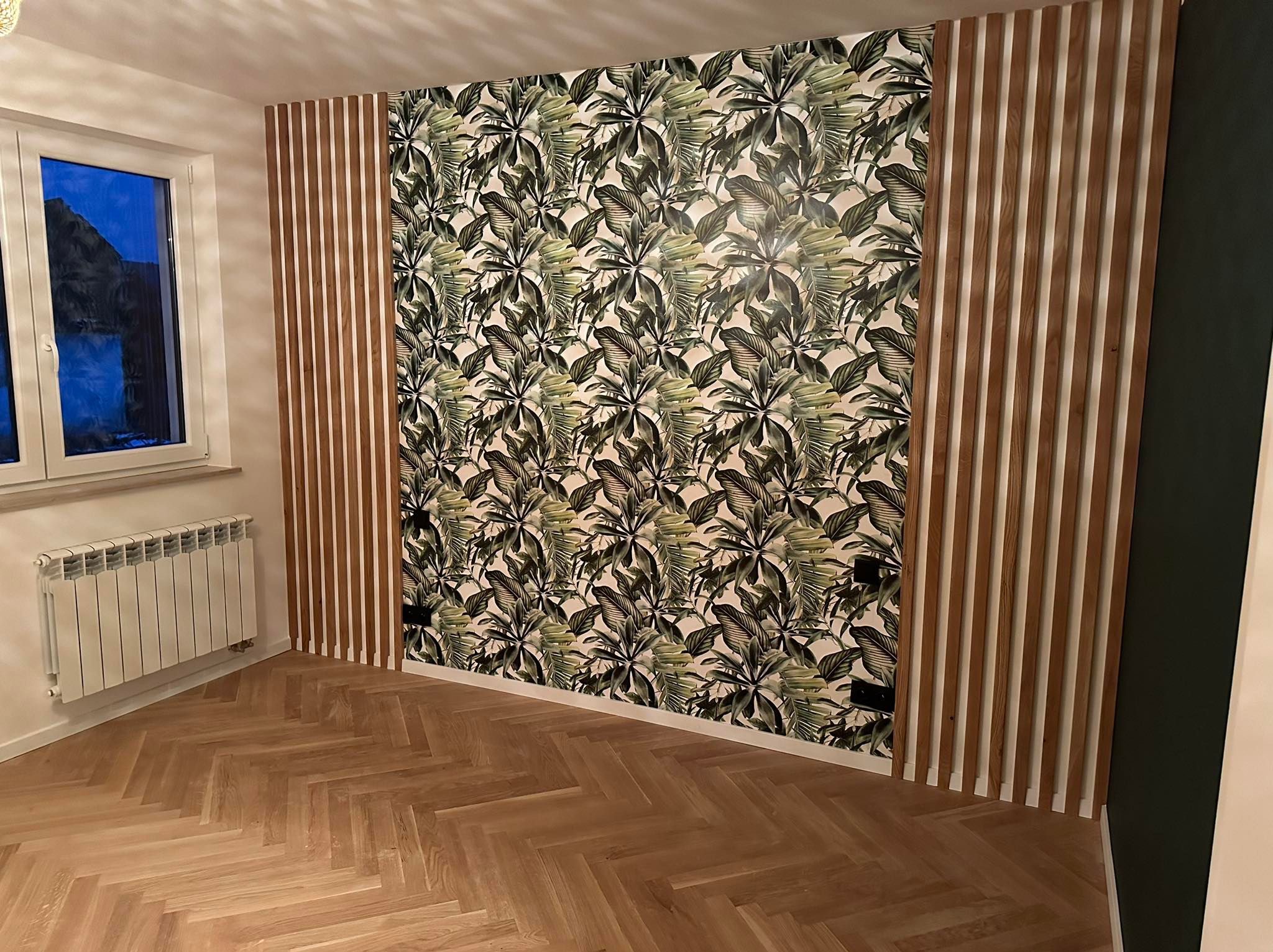 Wykończone wnętrze: parkiet w jodełkę, ściana z tapetą w tropikalne liście, drewniane lamele i grzejnik pod oknem. Nowoczesny design.