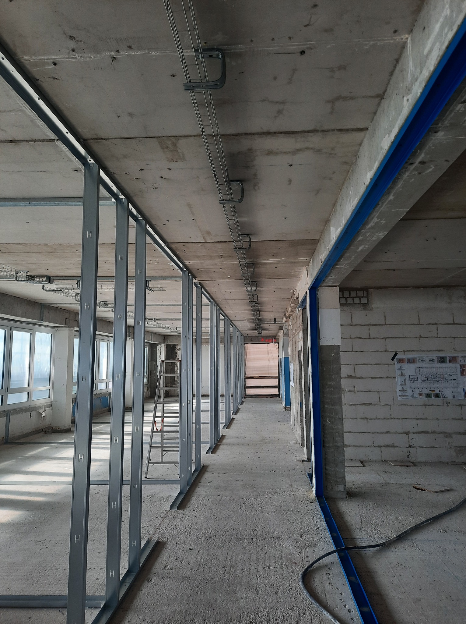 Metalowy szkielet ścian działowych w surowym wnętrzu, widoczne profile z oznaczeniami 'H', drabina i instalacje pod sufitem. W tle okna i plan budynku na ścianie.