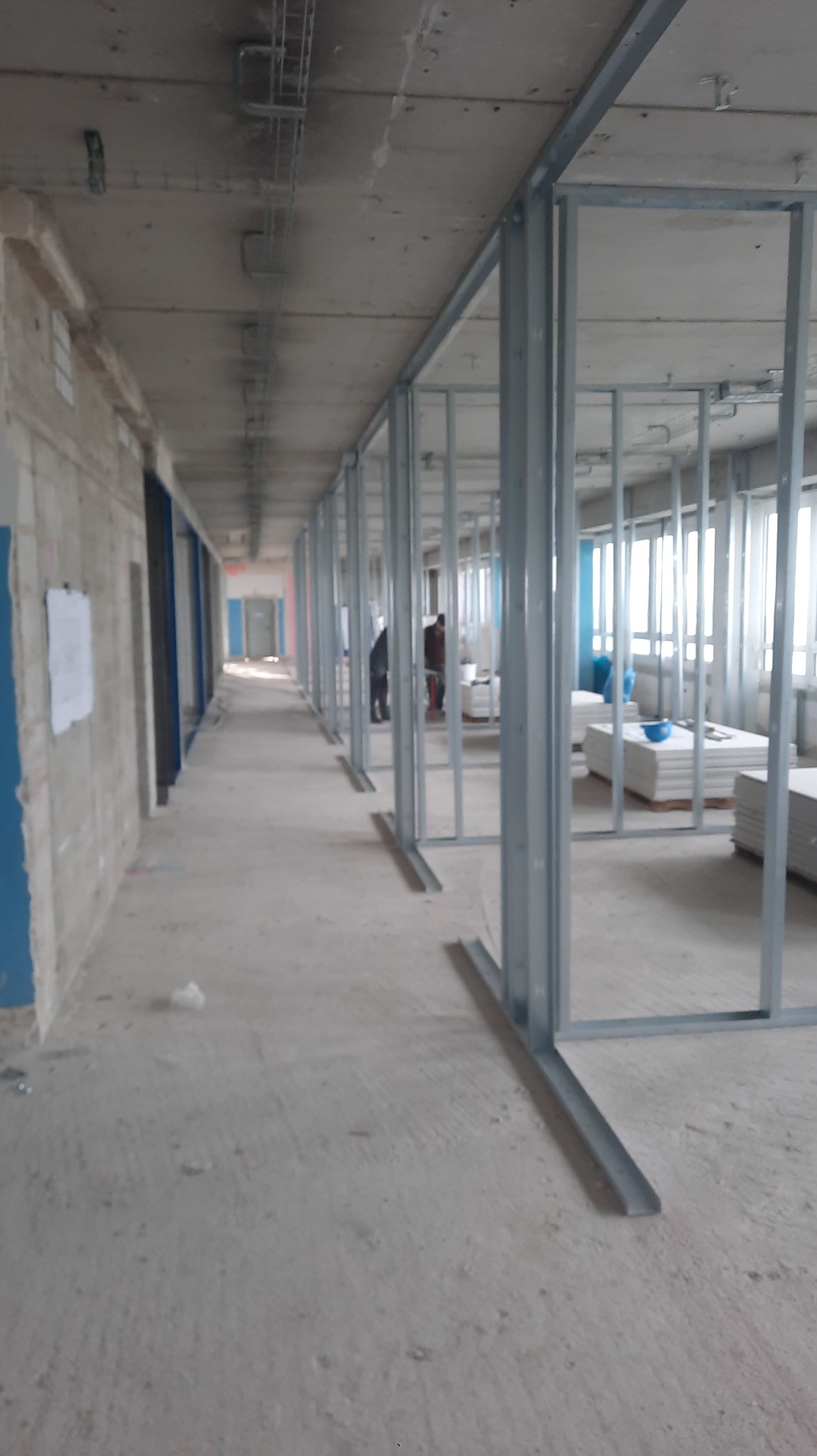 Montaż metalowej konstrukcji ścian działowych w długim korytarzu. Widoczne profile, płyty karton-gips i pracownicy. Surowy stan wykończenia.