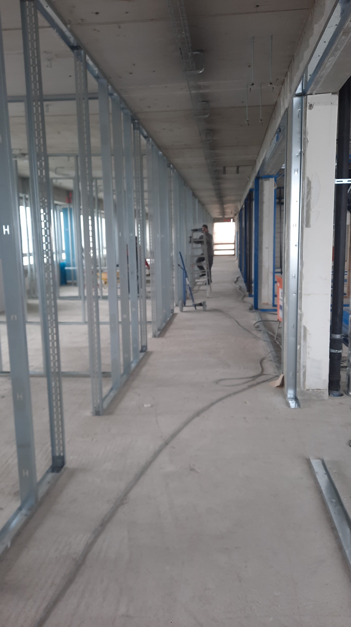Konstrukcja szkieletowa ścian działowych z profili stalowych w surowym wnętrzu, widoczne instalacje i drabina. Etap prac wykończeniowych w obiekcie komercyjnym.