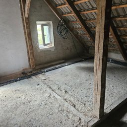 Krystian Rzempała - Instalacja przewodów elektrycznych w peszlach na poddaszu. Widoczne drewniane elementy konstrukcyjne dachu i okno. Przewody ułożone równolegle, przymocowane opaskami.