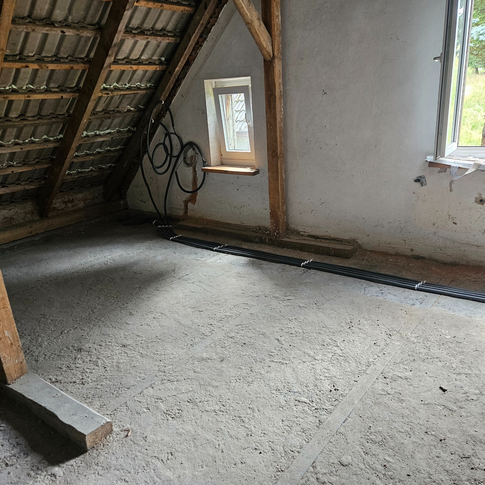 Instalacja przewodów w peszlu na poddaszu w surowym stanie. Widoczne okno, krokwie i podłoga w trakcie wylewki. Prace instalacyjne w nowym budynku.