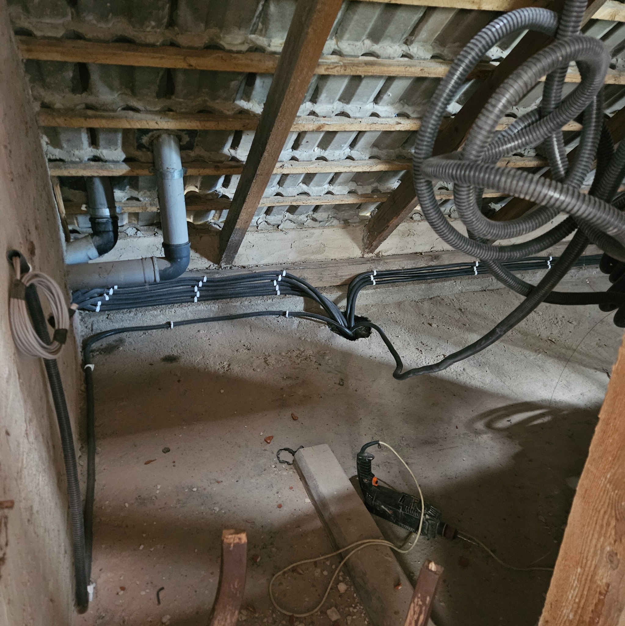 Instalacja kabli w peszlach, przymocowanych do ściany i sufitu. Widoczne rury i wiertarka na podłodze. Prace instalacyjne w trakcie realizacji.