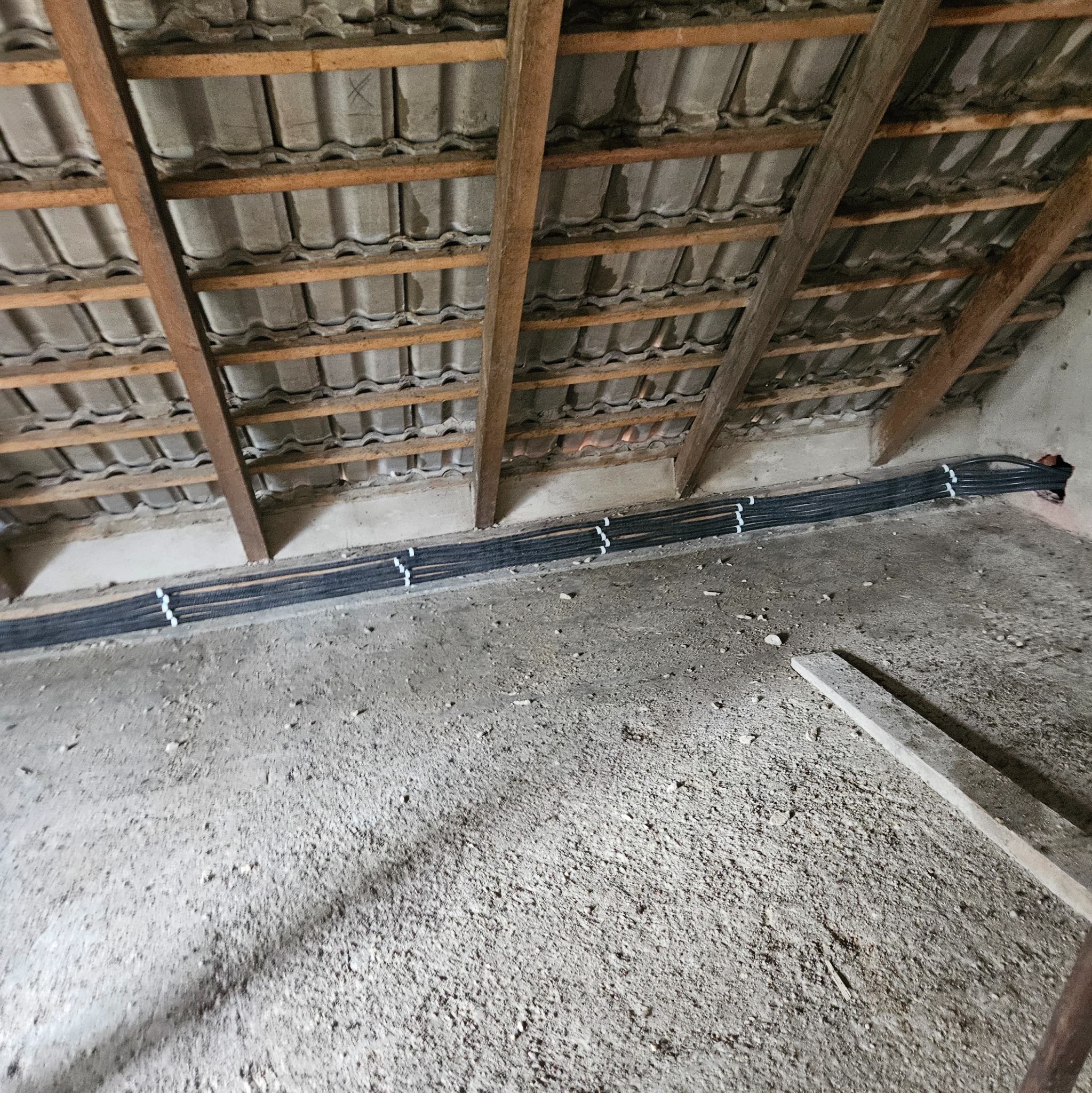 Kable elektryczne ułożone wzdłuż ściany poddasza z drewnianą konstrukcją dachu. Instalacja zabezpieczona białymi uchwytami. Surowy stan wykończenia.