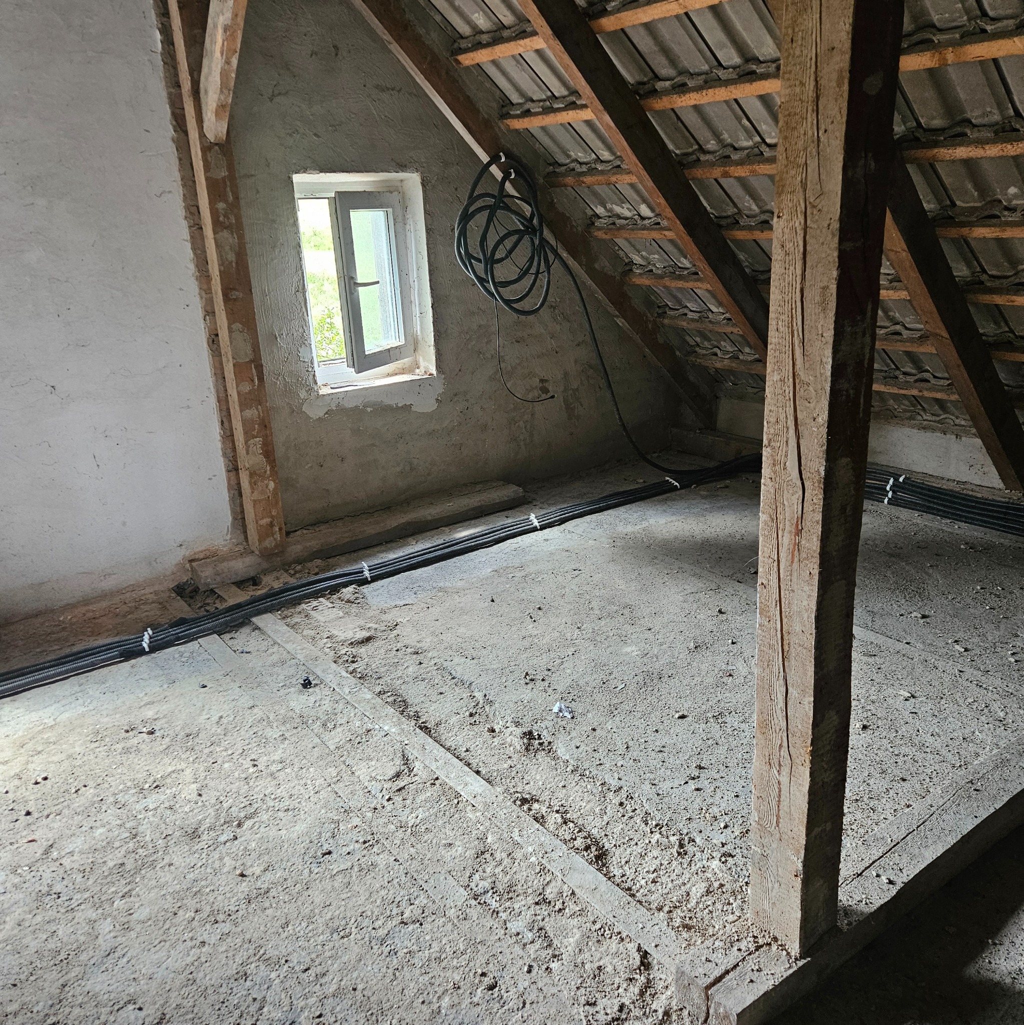 Instalacja przewodów elektrycznych w peszlach na poddaszu. Widoczne drewniane elementy konstrukcyjne dachu i okno. Przewody ułożone równolegle, przymocowane opaskami.