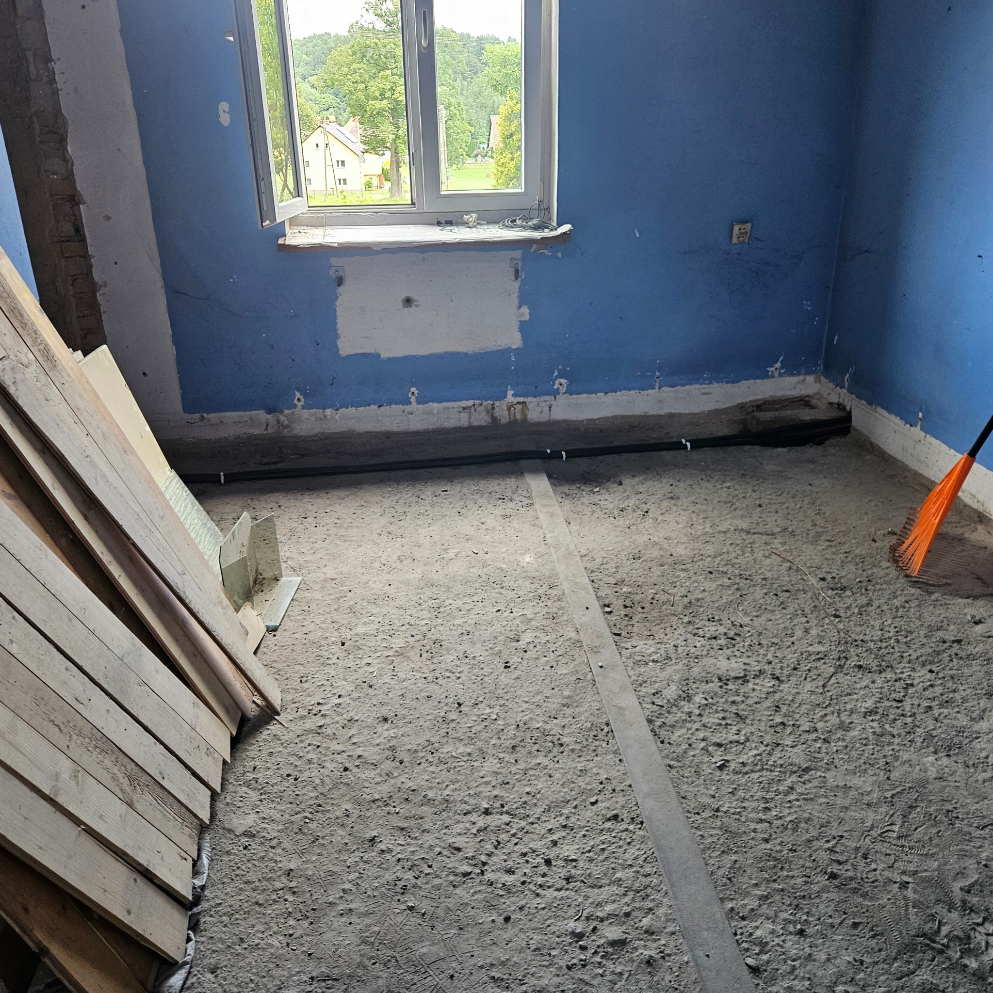 Wnętrze remontowanego pokoju z odsłoniętą instalacją elektryczną pod podłogą, widoczne deski i narzędzia, okno z widokiem na zieleń i niebieskie ściany.