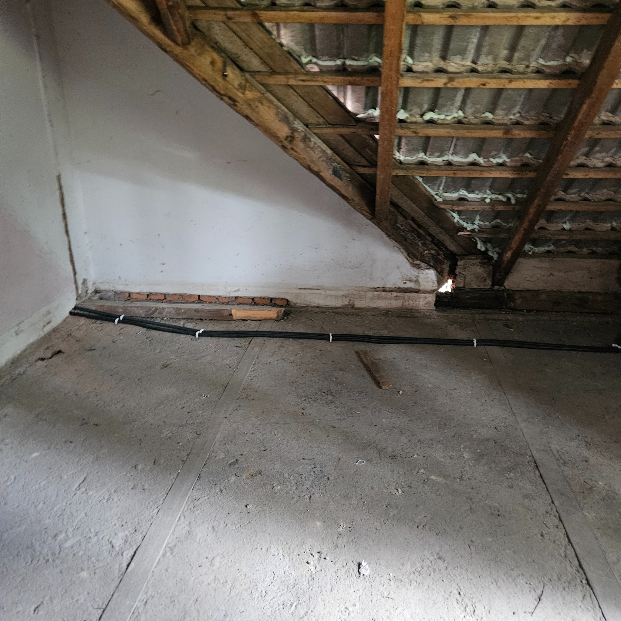 Kable w peszlu ułożone wzdłuż ściany na poddaszu w trakcie prac instalacyjnych. Widoczne elementy konstrukcji dachu i fragment ściany z cegłami.