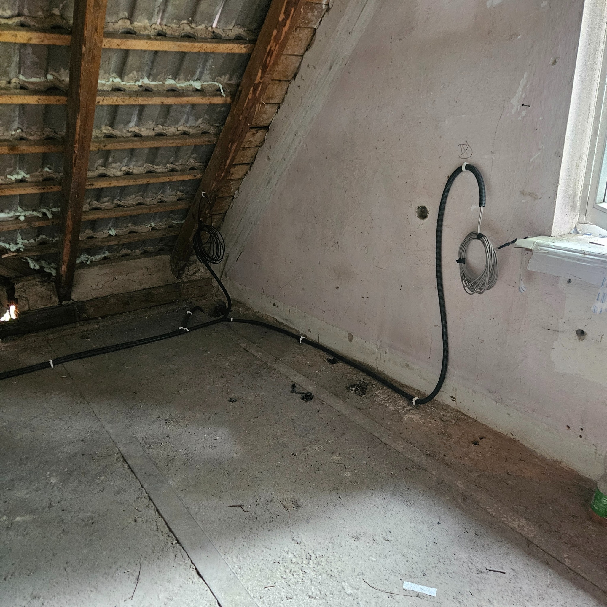 Instalacja przewodów w surowym wnętrzu poddasza. Widoczne kable przymocowane do ściany i podłogi, elementy konstrukcji dachu. Prace elektryczne w trakcie realizacji.