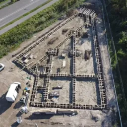 Widok z drona na drewniane szalunki fundamentów domu jednorodzinnego, z dwoma białymi samochodami dostawczymi obok placu budowy.