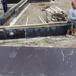 Sebastian Pągowski usługi budowlane SEBUD - Budowa fundamentów: szalunek zbrojony stalą, pracownik w kasku. Widoczne deski i narzędzia. Etap przygotowawczy fundamentów pod budynek.