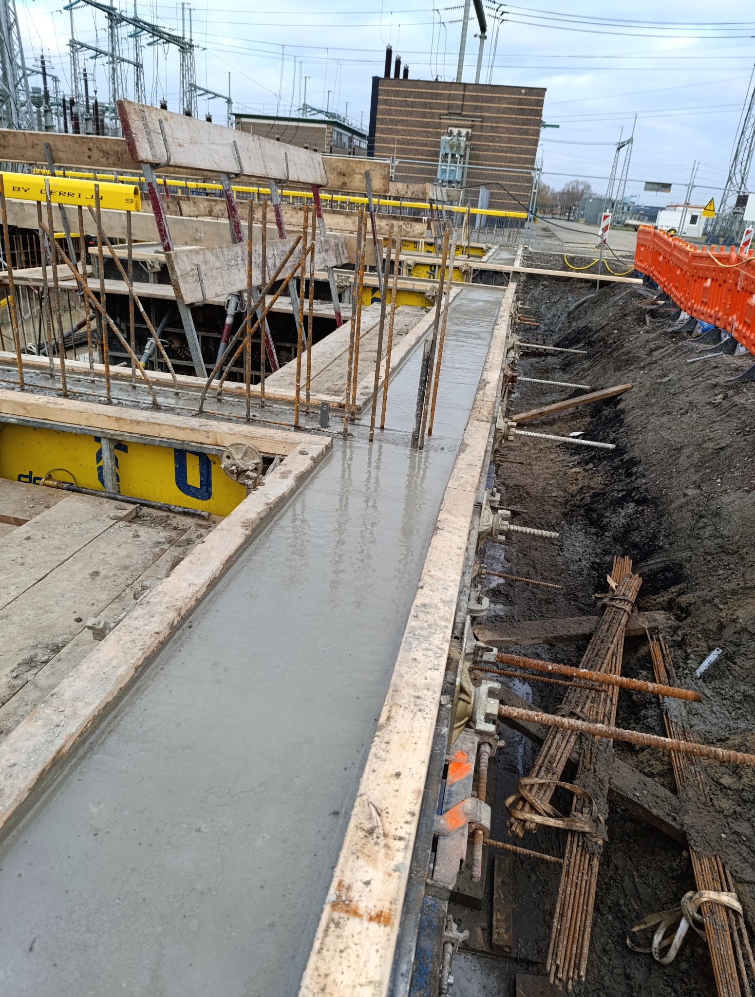 Wylany beton w szalunku z widocznym zbrojeniem, przygotowany do budowy fundamentów. W tle budynek stacji transformatorowej i słupy wysokiego napięcia.