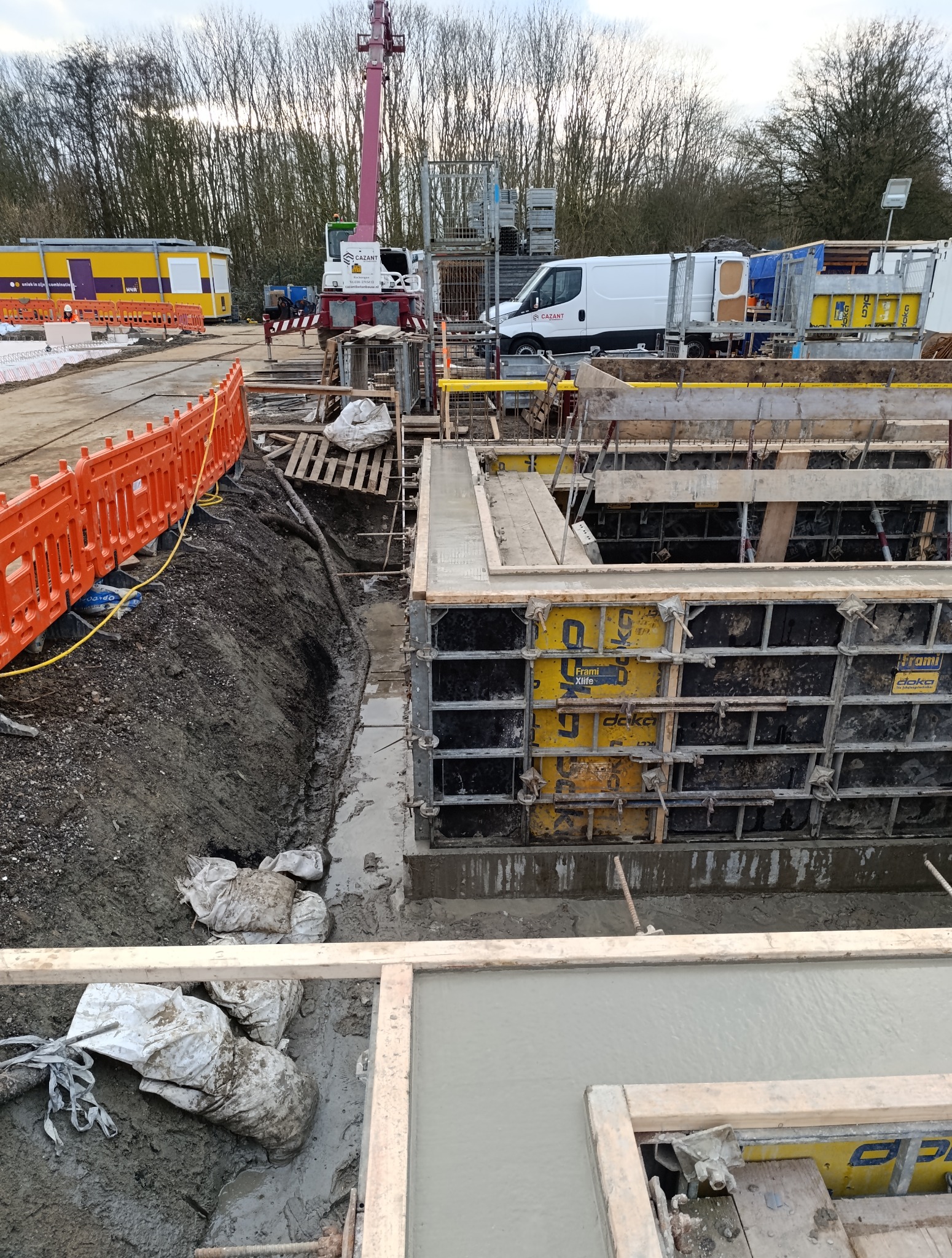 Wylane fundamenty z szalunkiem systemowym Doka, widok z góry. Na budowie obecny dźwig Cazant, zaplecze socjalne i pomarańczowe barierki. W tle drzewa.
