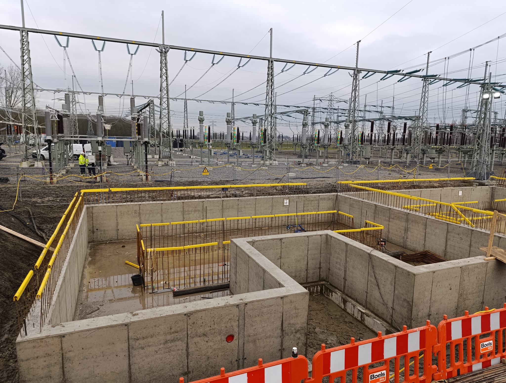 Wykop fundamentów pod stację transformatorową, zabezpieczony żółtymi barierkami. W tle słupy wysokiego napięcia i pracownicy w odblaskowych kamizelkach.